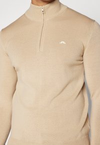 Beige ribbad stickad tröja med halvdragkedja och liten vit broderad logotyp på bröstet.