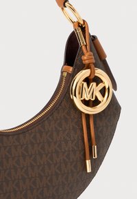 Braune monogrammierte Handtasche mit einem goldenen Logo-Anhänger, strukturiertem Material und einem Reißverschluss. Akzentuiert mit einem sandfarbenen Schultergurt und Beschlägen.