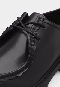 Mocassin en cuir noir avec bout arrondi, coutures détaillées et lacet en cuir noué en un nœud sur le coup de pied.