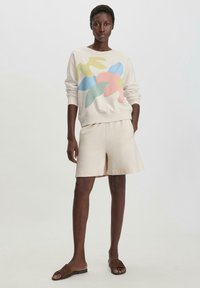 Lähe beige huppari, jossa on monivärinen abstrakti kukkapainatus; yhdistettynä beigeihin shortseihin ja ruskeisiin liukupohjakengiin. Sileä, pehmeä tekstuuri.