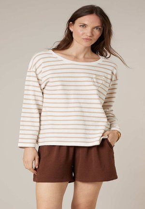 Femme portant un haut à manches longues rayé blanc et beige avec un motif cœur, associée à un short ample marron, debout avec une main dans la poche.