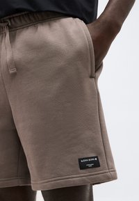 Κοντινό πλάνο ενός ατόμου που φορά καφέ σορτς Björn Borg με ελαστική μέση και κορδόνι, το χέρι ακουμπά στην τσέπη.