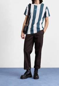 Pruhované tričko v tmavě modré a bílé, černé kalhoty a masivní černé boty. Na přední straně trička je viditelné malé logo.