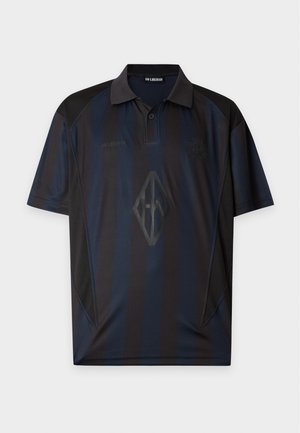 Homme portant un polo sombre et ample, un pantalon large en denim foncé et des chaussures noires, debout devant un fond clair uni.
