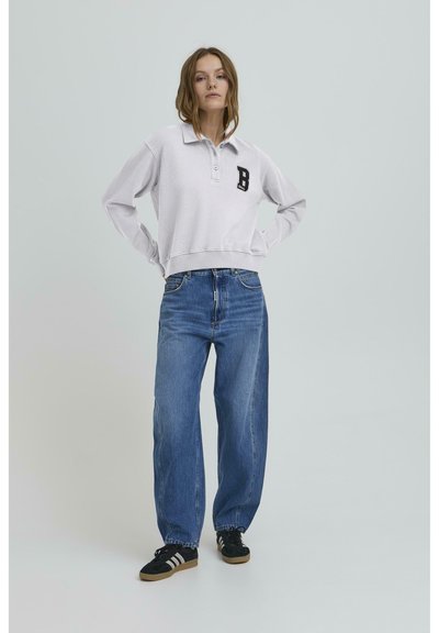 Svijetlo sivi dukserica s ovratnikom i crnim "B" logom na prsima, uparen s širokim plavim trapericama i crnim tenisicama.