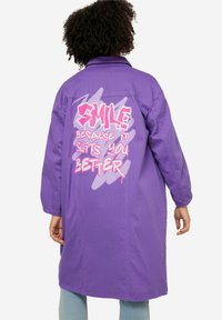 Lange paarse jas van katoen met een relaxed fit. Heeft een graffiti-achtige tekstontwerp in het roze op de achterkant.