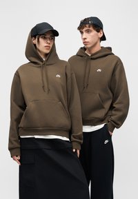 Dve osebi, ki nosita olivno zelena Nike kapucarja, črne kape in črne spodnje dele oblačil, stojita drug ob drugem na enostavnem sivem ozadju.