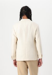 Beige op maat gemaakte blazer met een gelapte kraag, knoopmanchetten en textuurfabric. De achteraanzicht toont een strakke silhouet en een enkele vent.