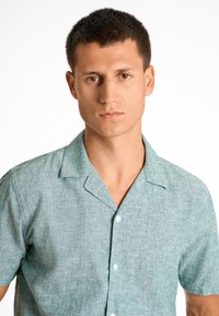 Korte mouw knoopshirt in lichtteal stof, geweven met een subtiel patroon. Heeft een kraag en witte knopen.
