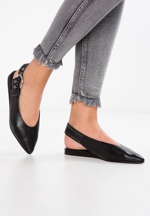 Pieds portant des ballerines slingback pointues noires avec des brides cloutées et un jean skinny gris à ourlet effiloché sur fond blanc.