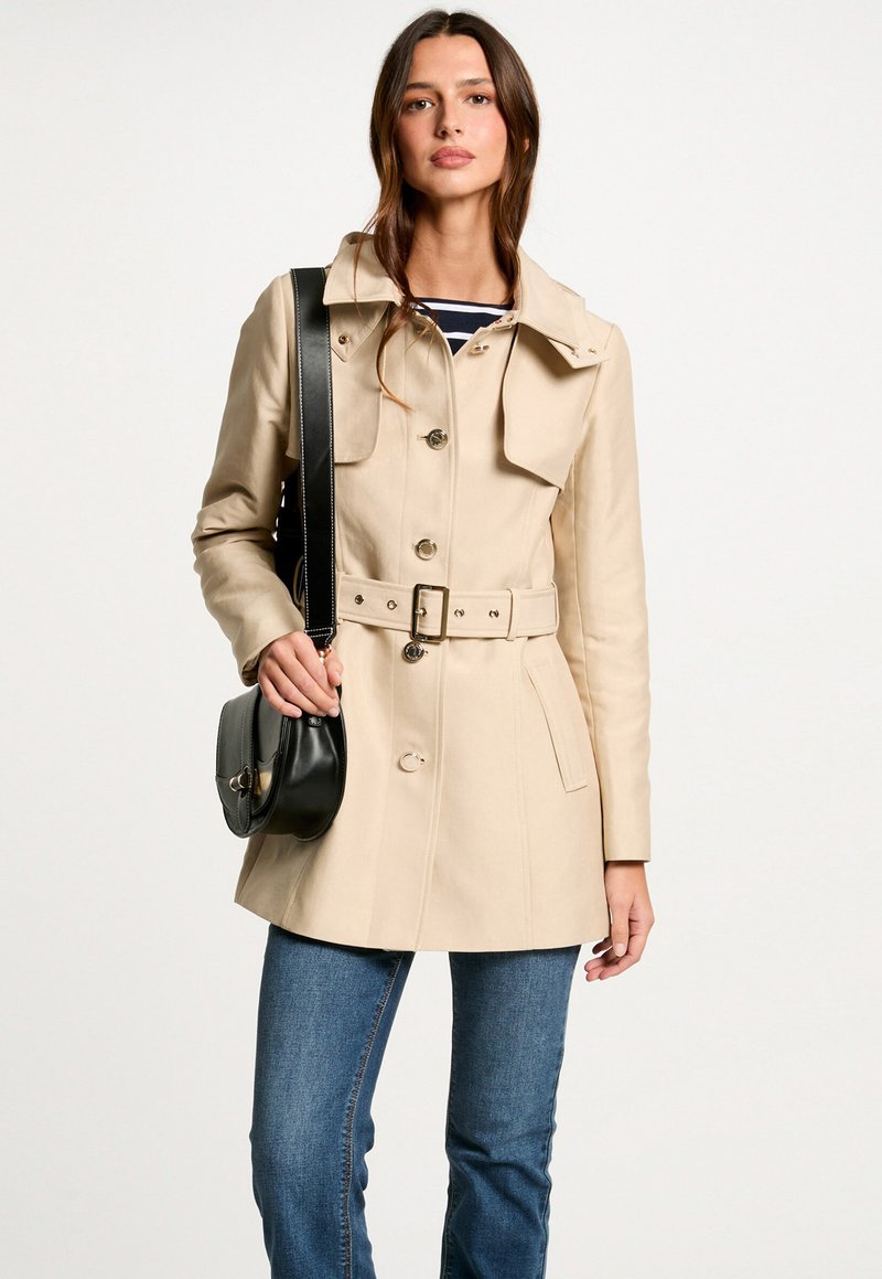 Beige trenchcoat met een ceintuur in de taille, dubbelzijdig knopen en grote kraag. G gedragen met donkere jeans en een zwarte schoudertas.