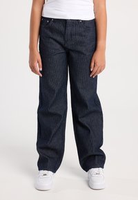 Lapsi, jolla on tummansiniset pinstripe-housut ja valkoiset tennarit, seisoo tasaisella valkoisella taustalla.