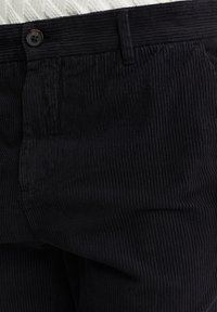 Detail černých manšestrových kalhot ukazuje pas s knoflíkovým zapínáním a poutky na opasek, zdůrazňující texturovanou tkaninu a švy.