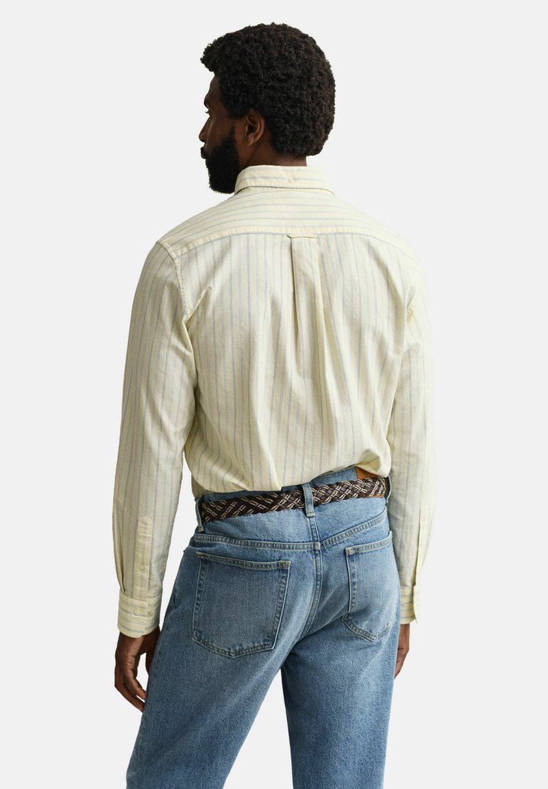 Hombre con cabello rizado y barba que lleva una camisa de manga larga a rayas de color amarillo claro, metida dentro de unos jeans azules con un cinturón tejido marrón, visto de espaldas.