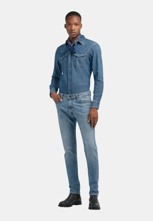 Hombre de pie con una camisa vaquera azul, vaqueros azul claro, botas negras y un pañuelo azul con estampado en el cuello, mirando hacia adelante con una mano en el bolsillo.