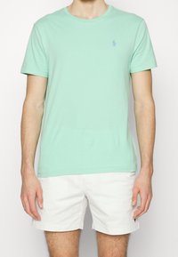 Lichtgroene katoenen t-shirt met een ronde halslijn en korte mouwen, met een klein blauw embleem op de linker borst. Gedragen met witte shorts.