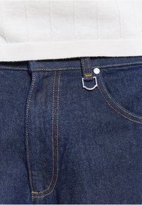 Pull en maille blanc rentré dans un jean en denim bleu foncé avec des coutures jaunes visibles et un porte-clés argenté à la taille.