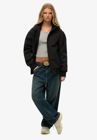 Sort, lynlåsløs jakke, grå tanktop og løse blå jeans med en stor guldspænde. Beige sneakers med et struktureret design.