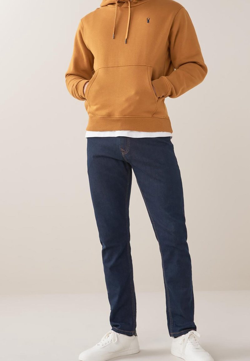 Personne portant un sweat à capuche moutarde avec une poche avant, un jean bleu foncé et des baskets blanches, debout devant un fond clair uni.