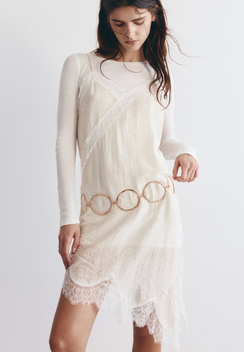 Femme portant un haut blanc à manches longues sous une robe caraco crème en dentelle avec une ceinture chaîne circulaire dorée, debout devant un fond uni.