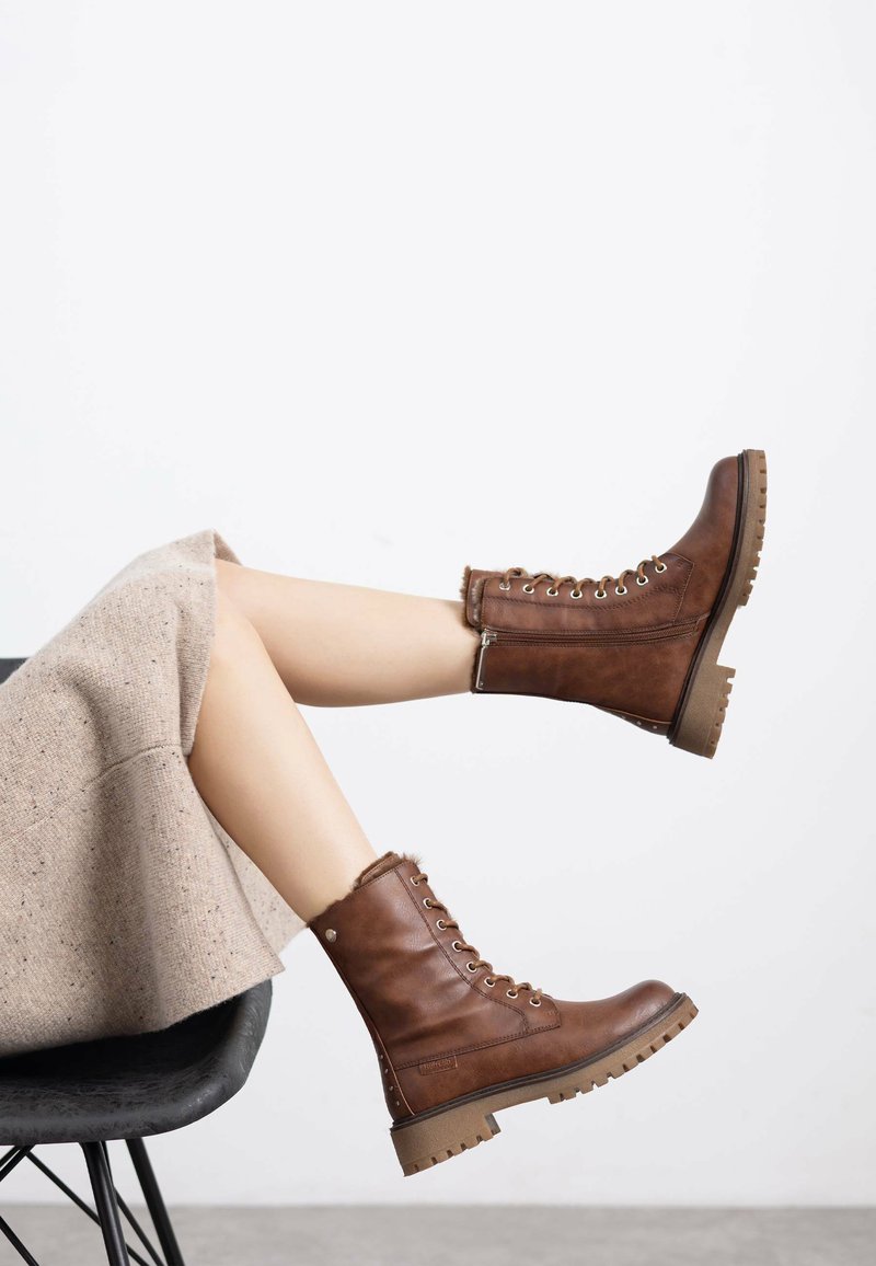 Botas de cuero marrón con cordones, hasta el tobillo, con una superficie texturizada, ojales y una suela de goma resistente. Un borde de pelo en la parte superior añade detalle.