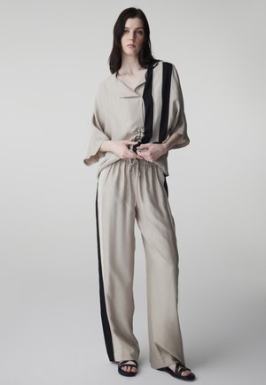 Mujer de pie vistiendo pantalones beige holgados y top a juego con rayas verticales negras, sosteniendo el dobladillo de la camisa con un fondo neutro.