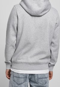 Grijze hoodie met een capuchon, geribde manchetten en onderkant. De stof ziet er zacht uit; gedragen over een wit shirt voor een gelaagde uitstraling. Jeans zijn zichtbaar aan de onderkant.