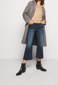 Abrigo largo a cuadros, suéter beige, jeans de pierna ancha azul oscuro, botas negras de tobillo con tacones cuadrados. Contrastes de materiales en toda la indumentaria.