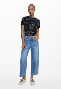 Zwarte t-shirt met witte cartoonafbeelding en tekst, gecombineerd met lichtblauwe wijde jeans met zwarte geborduurde accenten, en witte schoenen.