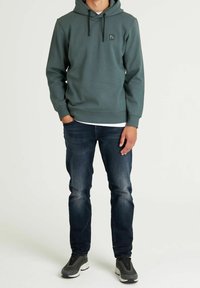 Felpa con cappuccio verde scuro, con tasca frontale e un piccolo logo. Indossata con jeans blu scuro e sneakers grigie. Tessuto liscio, vestibilità casual.