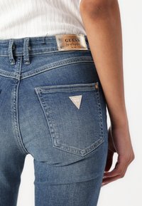 I jeans in denim blu presentano una vita media, cinque tasche e un piccolo logo triangolare sulla tasca posteriore. Il tessuto ha una texture classica, leggermente usurata.