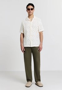Mann mit braunen Sonnenbrillen, kurzärmligem cremefarbenem Strickhemd mit Knöpfen, olivgrünen Hosen und beigen Schlupfschuhen, der vor einem weißen Hintergrund steht.