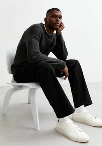 Maglione a maglia grigio scuro, pantaloni neri, sneakers bianche con polsini a costine, seduto su una sedia bianca contro uno sfondo chiaro.