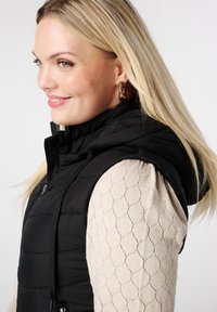 Zwarte gewatteerde vest met capuchon en rits, gedragen over een beige getextureerde lange mouwen top met een geometrisch patroon.