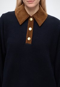 Femme portant un pull bleu marine avec un large col en velours côtelé marron et trois boutons dorés sur le devant.