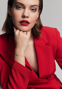 Czerwony dopasowany żakiet z głębokim dekoltem w kształcie litery V, złote kolczyki w kształcie serca oraz delikatna złota bransoletka z zawieszką w kształcie serca. Gładkie, wypolerowane tekstury.