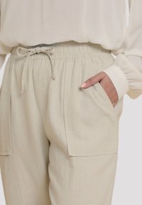 Ljust beige byxor med elastisk midja och dragsko, med sidfickor och en avslappnad passform, tillverkade av ett mjukt, texturerat tyg.