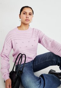 Pulover tricotat de culoare roz deschis, cu un design perforat, guler rotund și mâneci lungi, purtat cu blugi din denim albastru și o geantă neagră.