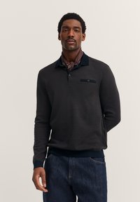 Dunkelgraues langärmeliges Polo mit Kragen, subtiler Struktur, Vordertasche und marineblauen Akzenten, kombiniert mit blauen Jeans.