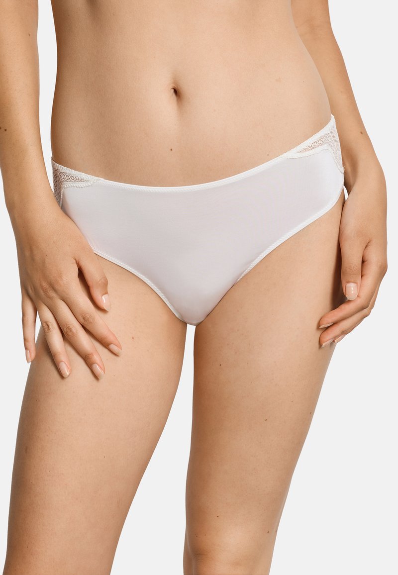 Frau trägt schlichte weiße Bikini-Unterwäsche mit Spitzendetails an den Hüften, steht mit locker an den Oberschenkeln hängenden Händen.