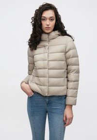 Béžová prešívaná bunda s vodorovným prešívaným vzorom, kapucňou a rukávmi strednej dĺžky. Nosená cez modré džínsy, s predným zipsom a vreckami.