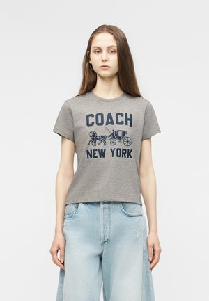 Młoda kobieta ubrana w szarą koszulkę z napisem "COACH New York" i grafiką konnej dorożki, w połączeniu z jasnoniebieskimi jeansami.