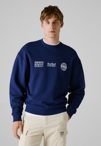 Sweatshirt bleu marine avec col rond, poignets et ourlet côtelés. Présente des logos blancs : "AUTHENTIC RACE GEAR" et "Red Bull Racing."