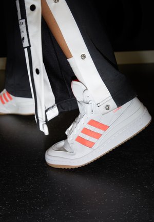 Baskets montantes blanches Adidas avec des bandes corail, portées avec un pantalon de survêtement noir et blanc à boutons-pression, sur un sol noir moucheté.
