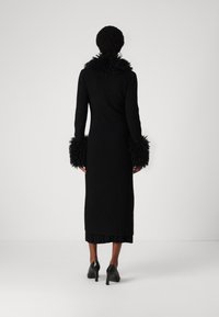 Zwarte geribbelde lange jurk met een geavanceerde, gefranjerde kraag en manchetten. Aansluitende silhouette, gecombineerd met zwarte hoge hakken.