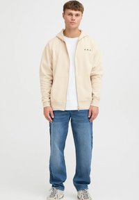 Beige Kapuzenpullover mit Reißverschluss, Kordelzugkapuze, Fronttaschen und schwarzem "A.R.C."-Schriftzug. Über einem weißen T-Shirt getragen und mit blauen Jeans kombiniert.