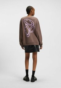 Sweatshirt brun à coupe oversize avec un design graphique violet au dos, associé à une jupe en cuir noir et des chaussures à plateforme.