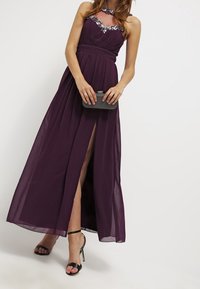 Kvinna i en golvlång ärmlös plommonfärgad klänning med dekorationer, som håller en liten svart clutchv äska och bär svarta högklackade sandaler.