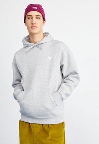 Grauer Hoodie mit verstellbaren Zugbändern und einer Fronttasche. Kombiniert mit senfgelben Cordhosen und einer lila Mütze.