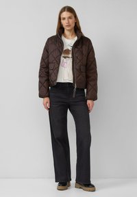 Brown quilted jacket with a high collar, zip closure, and front pockets. Worn over a light graphic t-shirt and paired with black jeans and chunky boots.
